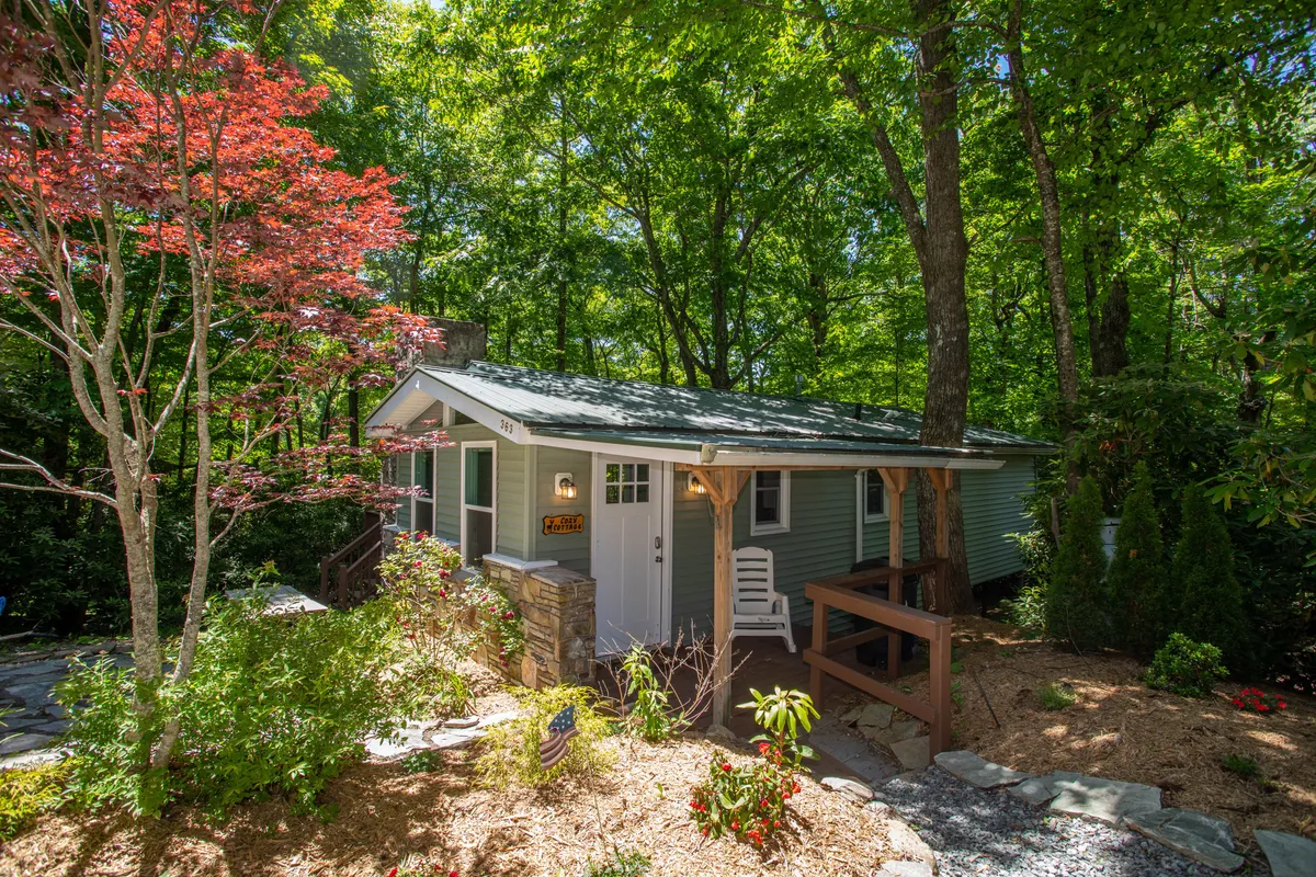 The image shows a charming small cottage nestled among trees, with a striking red tree providing a burst of color beside the house. A stone pathway leads up to a welcoming front porch, where cozy seating invites guests to relax and take in the tranquil surroundings. The scene feels peaceful, secluded, and perfect for unwinding in nature.