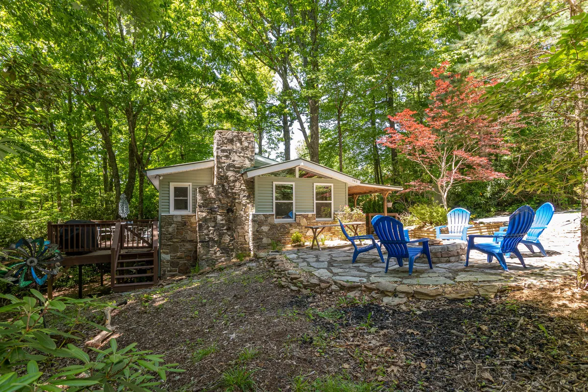 An inviting outdoor setting featuring a circular stone fireplace, blue Adirondack chairs, and lush greenery, set on a stone patio. The scene suggests tranquil outdoor gatherings and relaxed socializing, with ample space for groups to unwind.