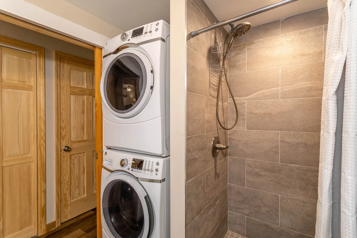 A practical and welcoming bathroom scene, featuring a stacked washer and dryer, a sleek stone-tiled shower area, and wooden doors. The overall look is modern and clean, emphasizing convenience and relaxation.