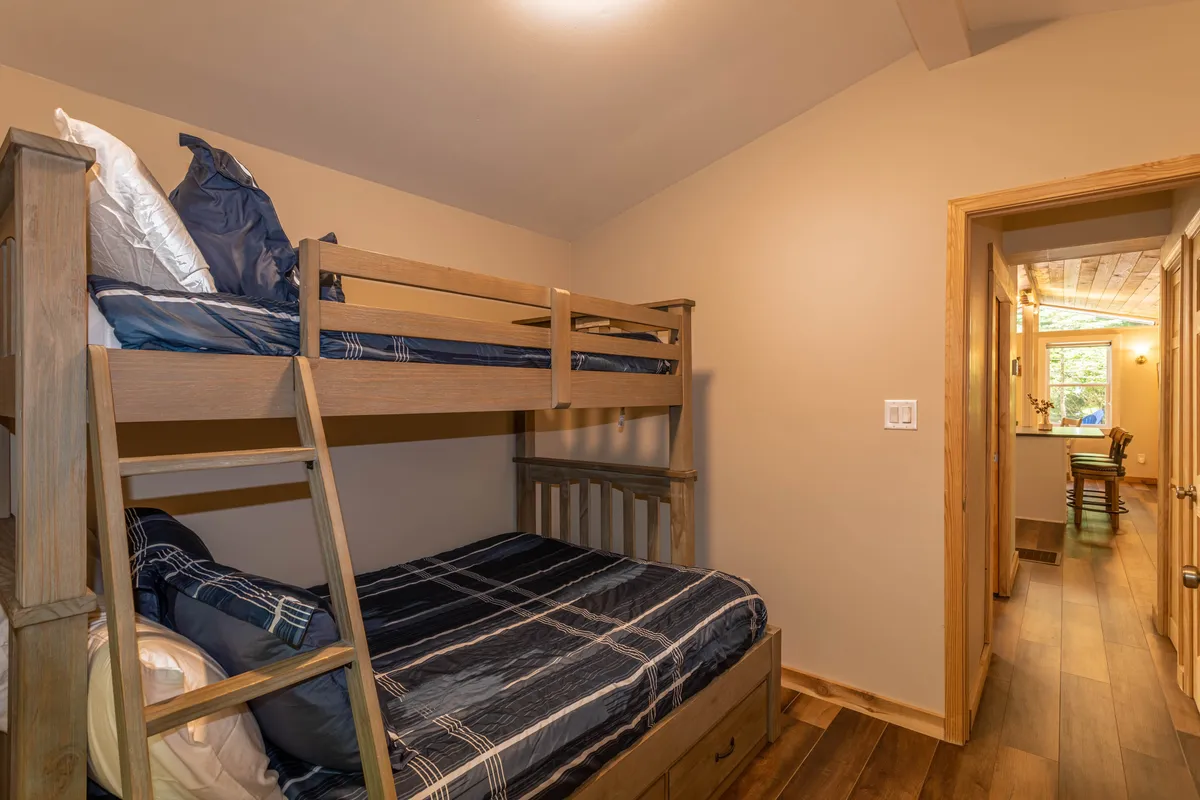 This image features a cozy bedroom with bunk beds, wooden flooring, and a doorway leading toward the kitchen. The space feels fun and welcoming, evoking memories of kids playing games or sharing stories, and highlights the home’s family-friendly sleeping arrangements.