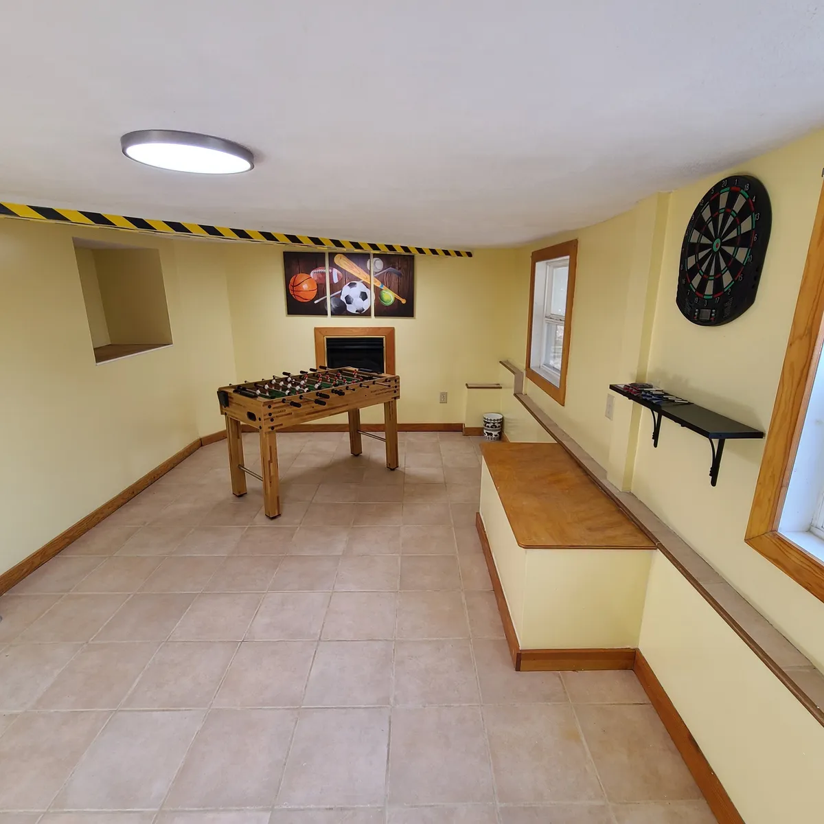 The image showcases a vibrant game room featuring a foosball table in the foreground, a kid-friendly dartboard mounted on the wall, and sports-themed artwork that adds playful energy to the space. The lighting and furnishings evoke a relaxed, fun-filled atmosphere ideal for family competitions and group activities.