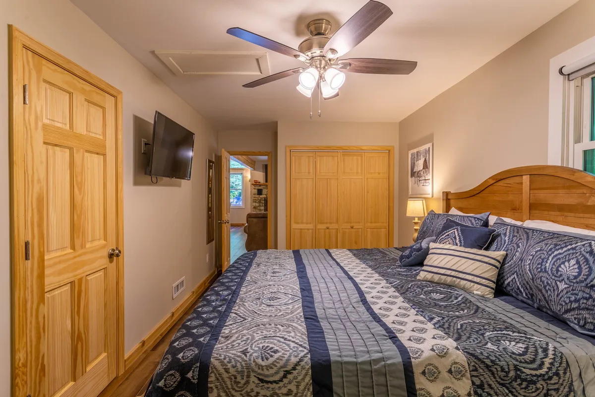 This image shows a spacious bedroom with a plush king bed adorned with decorative pillows, wooden furniture, a ceiling fan with light, and a wall-mounted Smart TV. Morning light streams softly through the windows, creating an inviting and restful atmosphere.