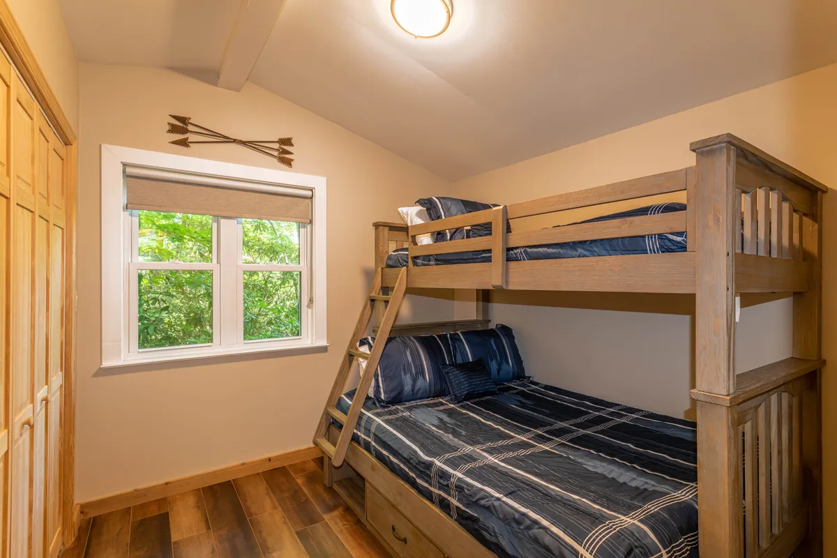 This image highlights a cozy bedroom with a twin-over-full bunk bed positioned near a window, a solid wooden wardrobe, and soft bedding. The space feels playful yet restful, ideal for children or friends, and offers a glimpse of nature outside the window.
