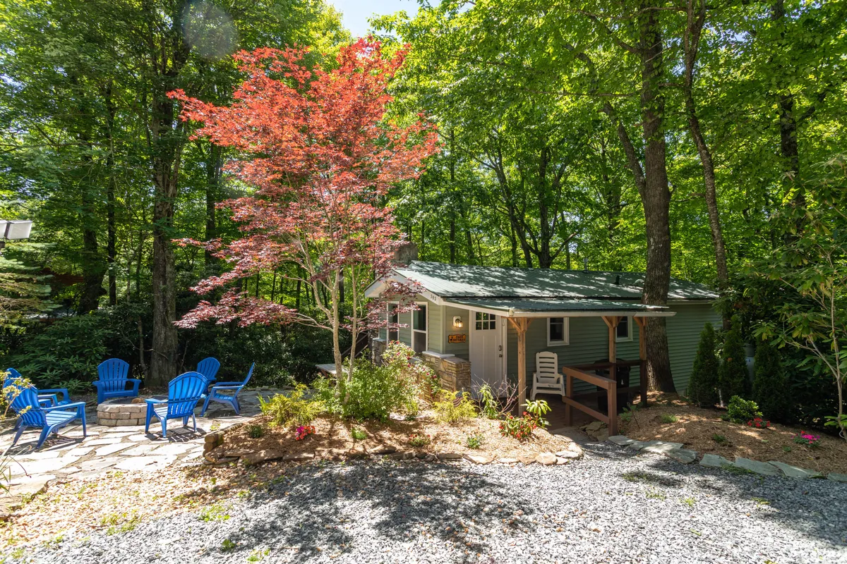 A tranquil exterior shot featuring the cottage, blue Adirondack chairs, a stone pathway, and a vibrant red maple tree. The setting emphasizes peaceful relaxation and the inviting charm of the property’s outdoor spaces.