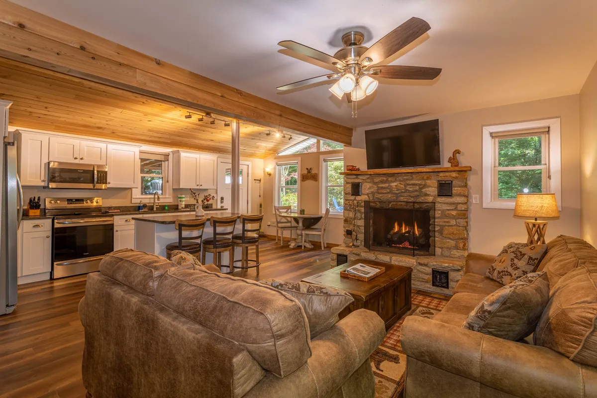 This inviting open-concept space features a stone fireplace as its centerpiece, flanked by plush, comfortable sofas and large windows that bathe the living area in natural light. The kitchen island is visible in the background, seamlessly connecting the living, dining, and kitchen zones. The overall atmosphere is warm, welcoming, and perfect for gatherings or cozy evenings at home.