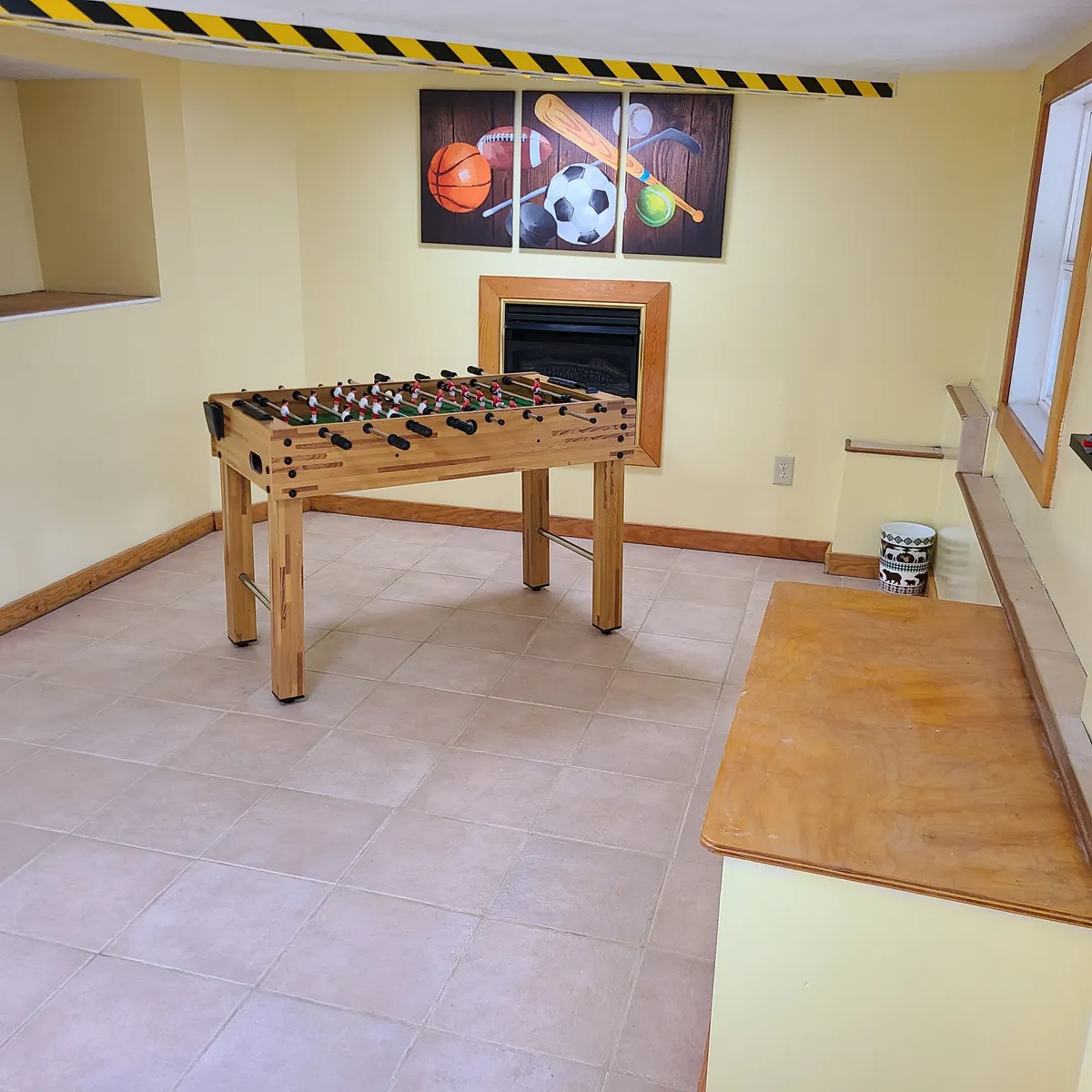 This inviting interior image centers around a foosball table surrounded by playful, sports-themed artwork and a fireplace. The arrangement suggests an inviting space for friendly competition, laughter, and quality time with loved ones.