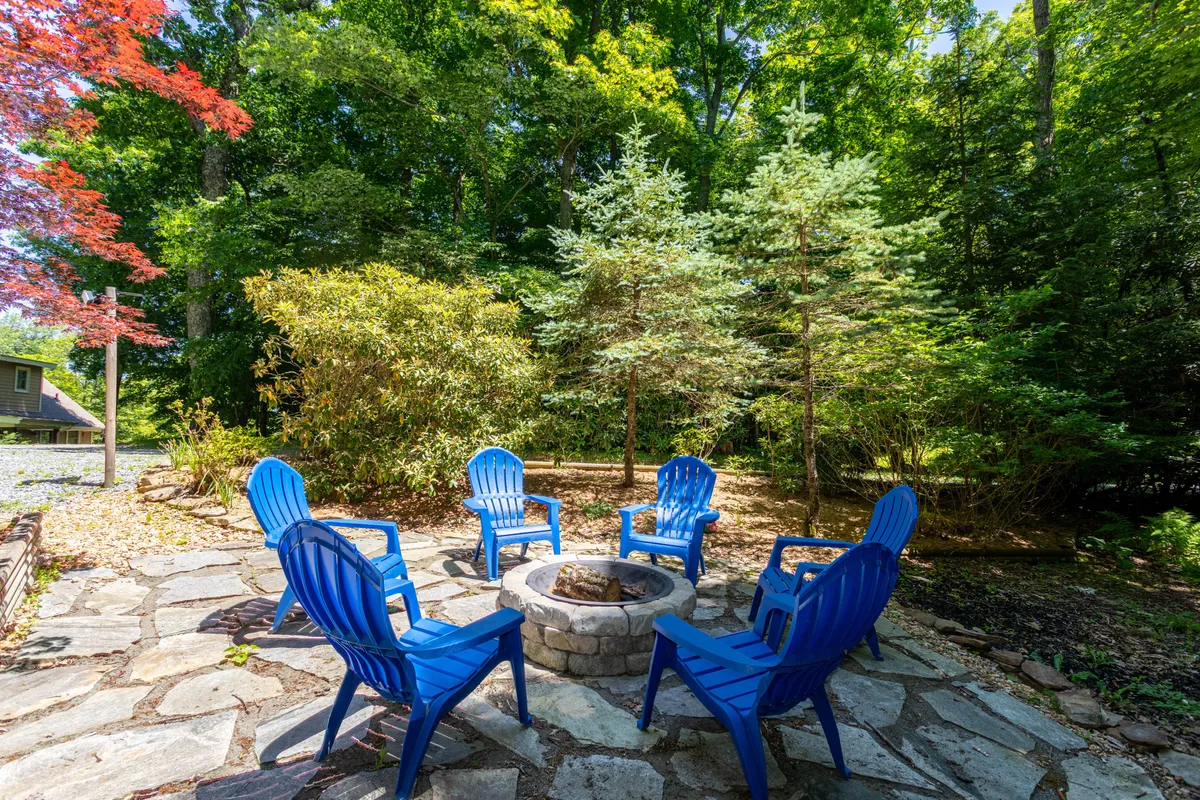 A cozy stone fire pit surrounded by blue Adirondack chairs, set amidst vibrant greenery. The scene suggests warmth, gathering, and the classic mountain experience of sharing stories and roasting marshmallows by the fire.