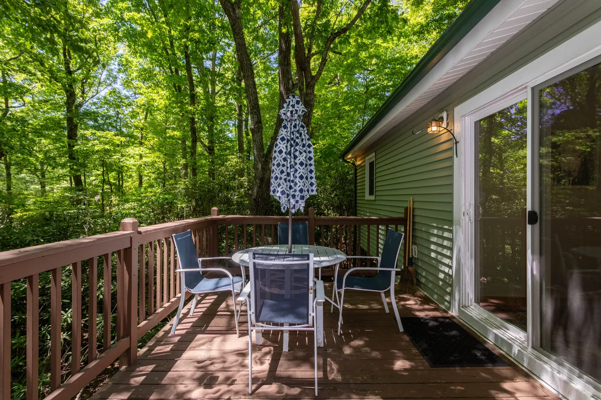 An exterior view of a patio area with a table and chairs shaded by an umbrella, surrounded by lush greenery. The layout offers an inviting spot for meals, reading, or conversation, with easy access for all guests.